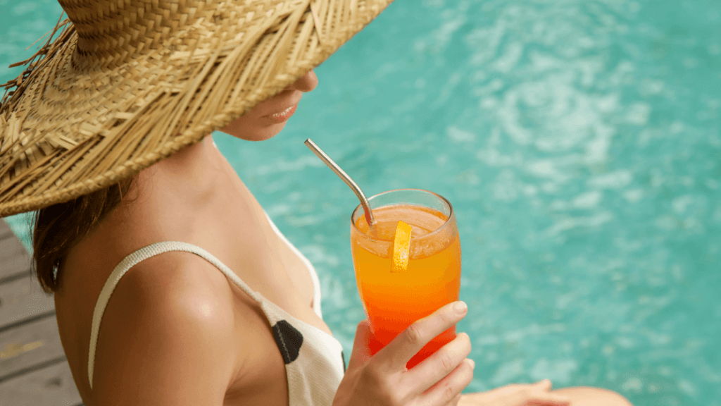 Person in a sunhat with their feet in the water drinking an orange cocktail.