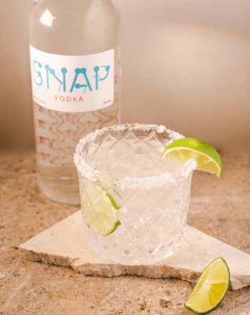 Clear cocktail in a salt-rimmed glass with a lime wedge on the rim and a slice of lime in the glass with a bottle of SNAP Vodka in the background..