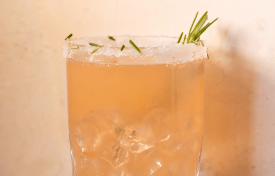A peach-colored cocktail in a glass with sprigs of rosemary sticking out of the glass.
