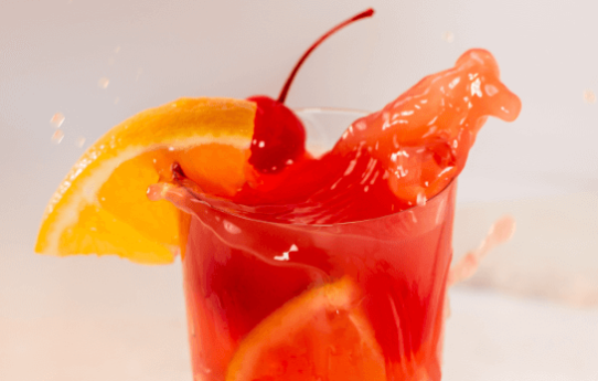 A glass with a red cocktail splashing out with orange slices and maraschino cherries in and around the glass.