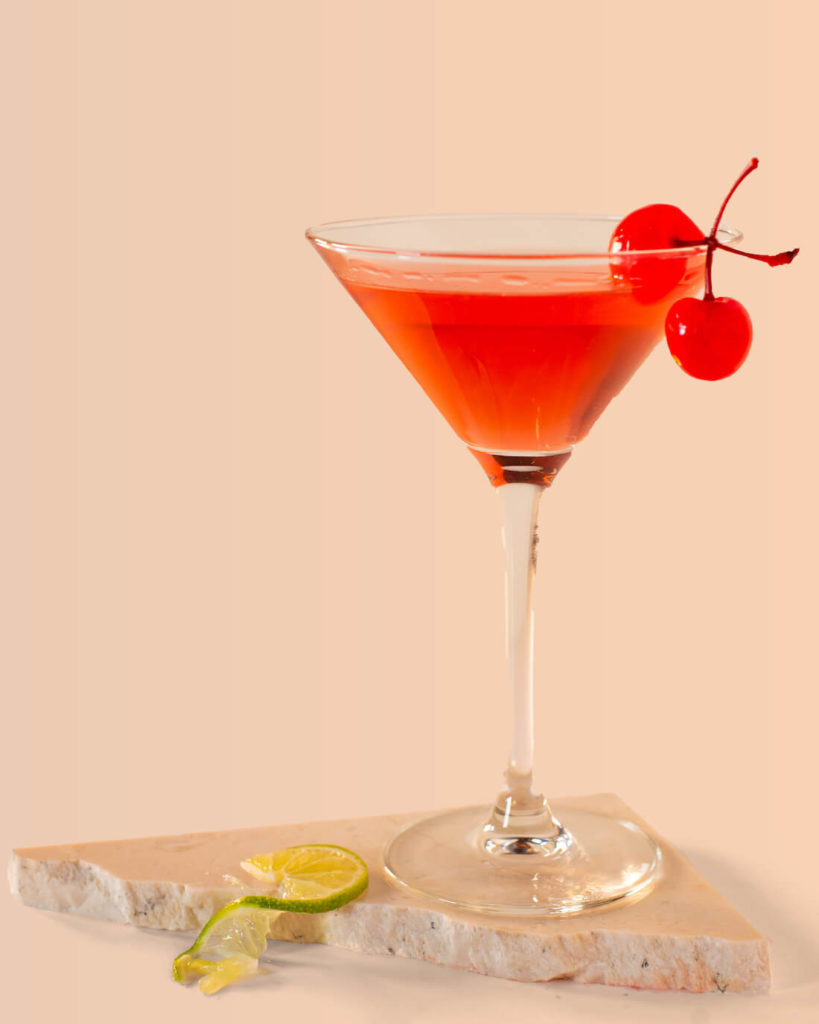 Red cocktail in a martini glass with maraschino cherries on the rim.